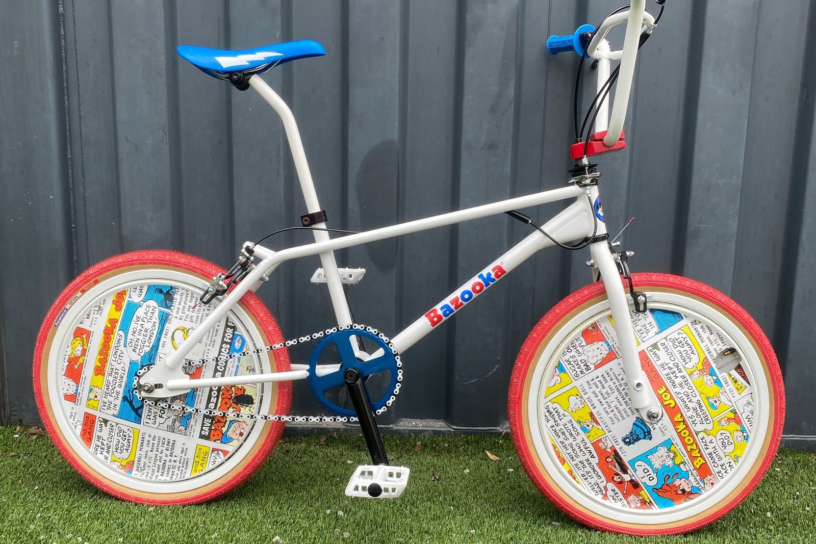 A white bike with blue handlebars, gear and seat and the Bazooka logo on the frame, featuring red wheels with Bazooka Joe comics in the center. The bike is on grass and leaning against a gray fence.
