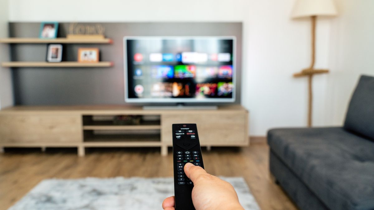 A hand holds up a remote in front of a television screen.