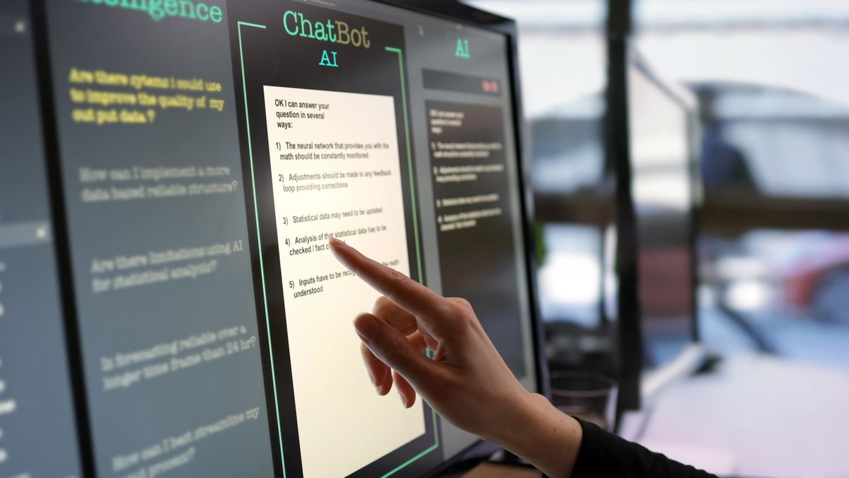 Close-up stock photograph showing a touchscreen monitor being used in an open plan office. A woman’s hand is asking an AI chatbot pre-typed questions & the Artificial Intelligence website is answering