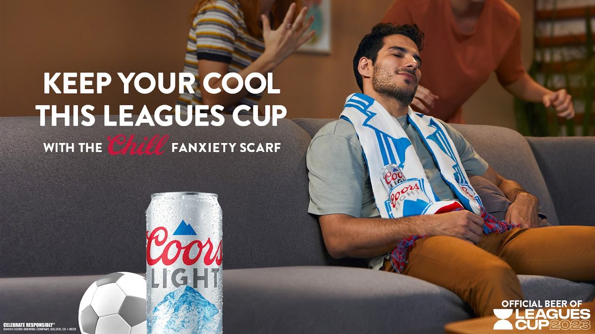 A man sits on a couch wearing a Coors Light branded soccer scarf