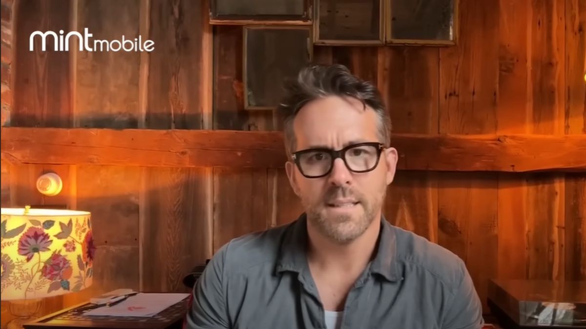 Actor Ryan Reynolds, wearing a gray button-up and glasses, is seated in front of wooden walls, with a Mint Mobile logo in the top left corner of the image.