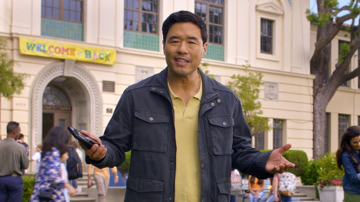 Actor Randall Park stars in Amazon’s back-to-school campaign and is seen standing, wearing a dark jacket and holding a phone, in front of a school.