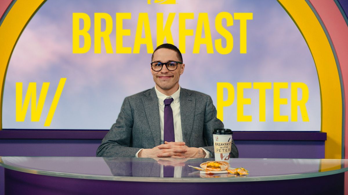 Comedian Pete Davidson, wearing a dark suit and tie and glasses, sits behind a desk as a morning show host