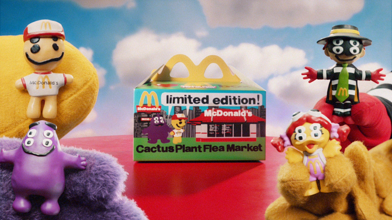 Four McDonald's figurines designed by Cactus Plant Flea Market encircle a Happy Meal placed on a red table against a sky-blue backdrop.